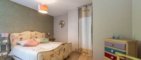 A bedroom with a floral bedspread, a bedside lamp, a wooden dresser, and a hanging light fixture.