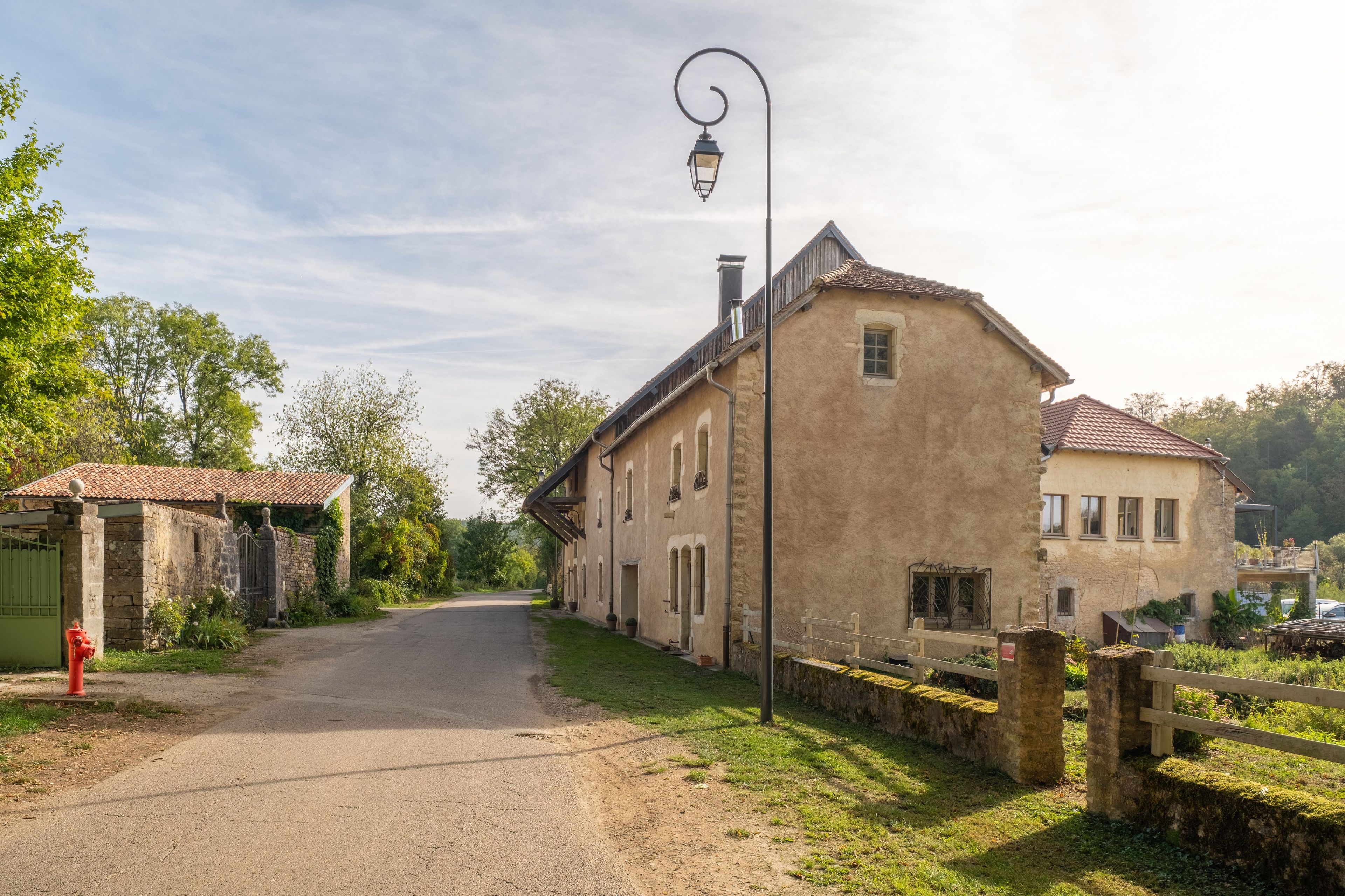 A rural road leading to a traditional stone building with a tiled roof, a street lamp, and a green gate.