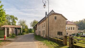 A rural road leading to a traditional stone building with a tiled roof, a street lamp, and a green gate.