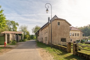 A rural road leading to a traditional stone building with a tiled roof, a street lamp, and a green gate.