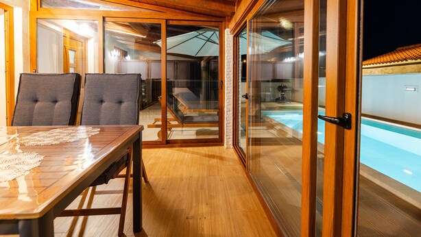A wooden deck with a dining table and chairs, a glass-enclosed sleeping area, and a view of an outdoor swimming pool. - Holiday Home Mareta with pool (Privlaka)