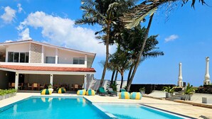 Une piscine avec des chaises longues, un bâtiment doté d’un toit rouge, des palmiers et un ciel dégagé.