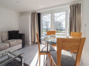 A modern living room with a glass dining table, wooden chairs, a sofa, and a view through large windows. - Beacon Fell Cottage 2 (Preston)