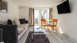 A living room with a sofa, a glass coffee table, a dining table with chairs, a TV mounted on the wall, and a large window with curtains. - Beacon Fell Cottage 2 (Preston)
