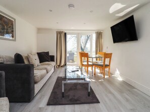A living room with a sofa, a glass coffee table, a dining table with chairs, a TV mounted on the wall, and a large window with curtains. - Beacon Fell Cottage 2 (Preston)