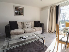A modern living room with a glass coffee table, a sofa with patterned cushions, a framed picture on the wall, and a dining area with a glass table and chairs. - Beacon Fell Cottage 2 (Preston)