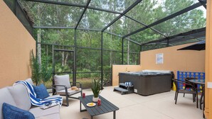 A covered patio with a sofa, chairs, a table, a hot tub, and a view of the forest. - Walt's Disney Villa | Hot Tub & NO Resort Fees (Kissimmee)