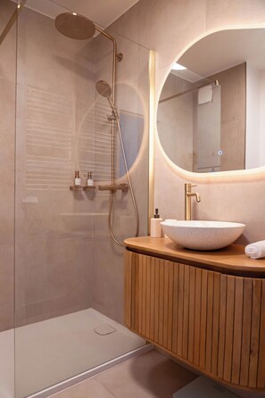 Ein modernes Badezimmer mit einer gläsernen Duschkabine, einem hölzernen Waschtisch mit rundem Spiegel und einem weißen Waschbecken.