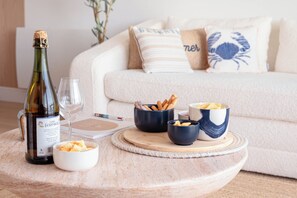 Eine Flasche Wein, Gläser und Schüsseln mit Snacks auf einem runden Tisch, im Hintergrund ein weißes Sofa.