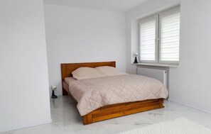 Une chambre avec un lit en bois, des murs blancs, une fenêtre avec des stores et un radiateur.