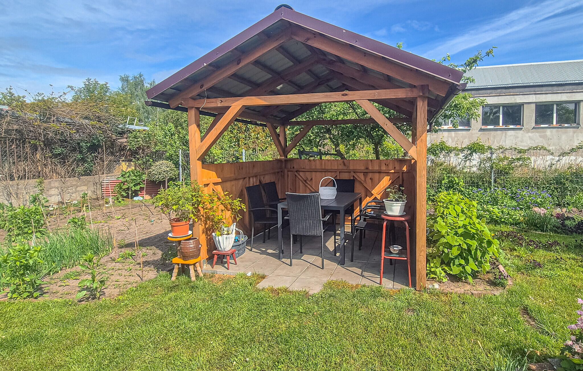 Een houten pergola met een tafel en stoelen, omringd door plantenpotten en een tuin.