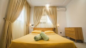 A neatly made bed with a yellow bedspread, a wooden dresser, and a wall-mounted air conditioner. - Villa Rosette, Calme, confort, services premiums (Lomé)