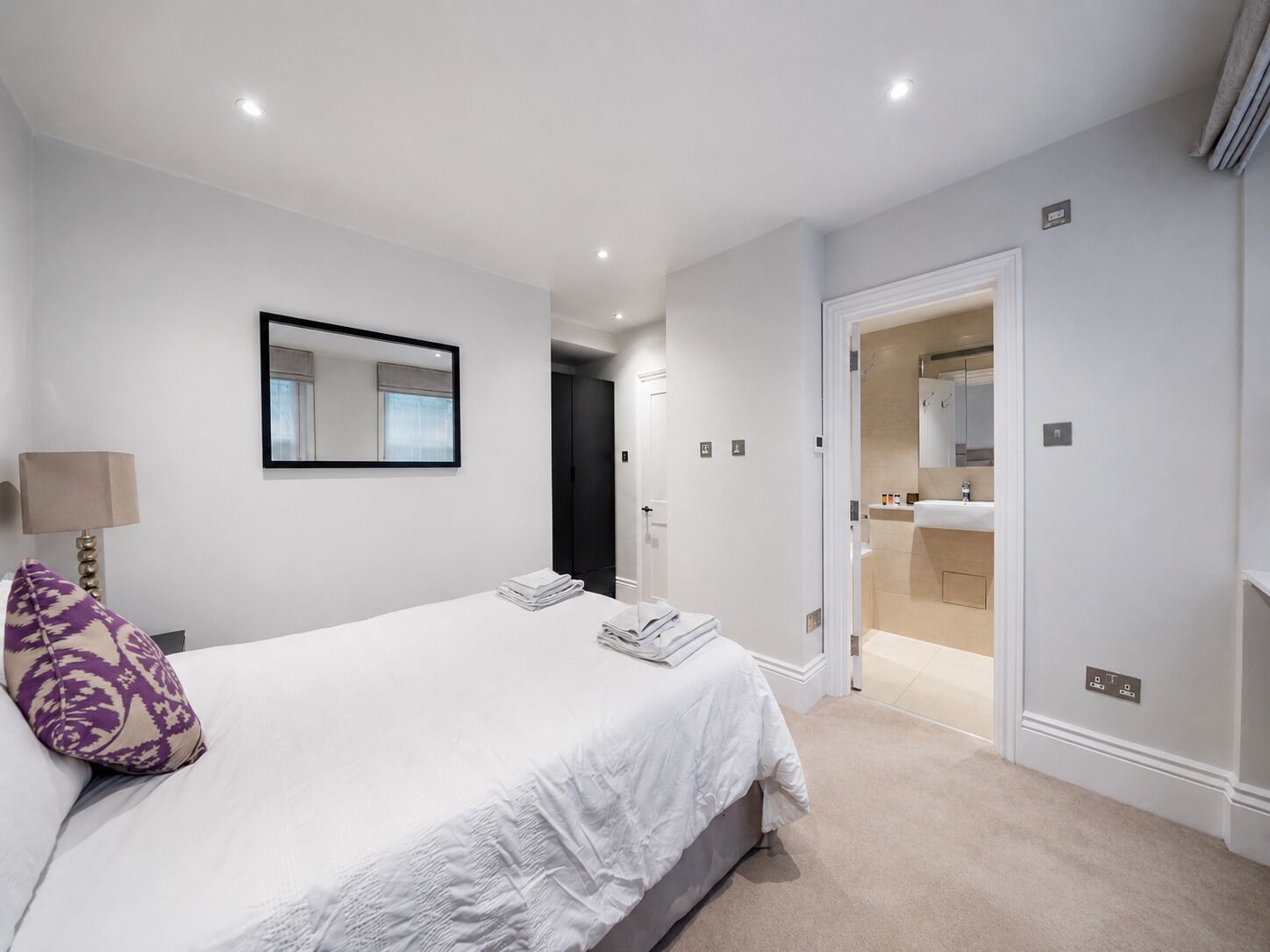 A bedroom with a bed, a bedside lamp, a mirror, and a door leading to a bathroom.