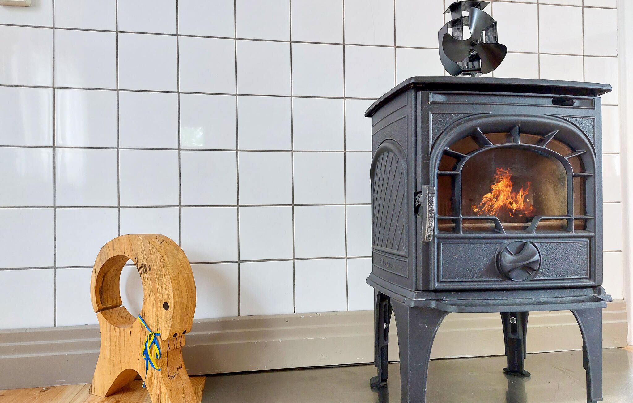 Ein Kaminofen mit Feuer darin, ein Holzschemel mit blauem Band und eine geflieste Wand im Hintergrund.