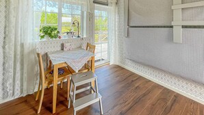 Et værelse med et træbord, stole, en lille skridestige og et vindue med udsigt til grøn vegetation.