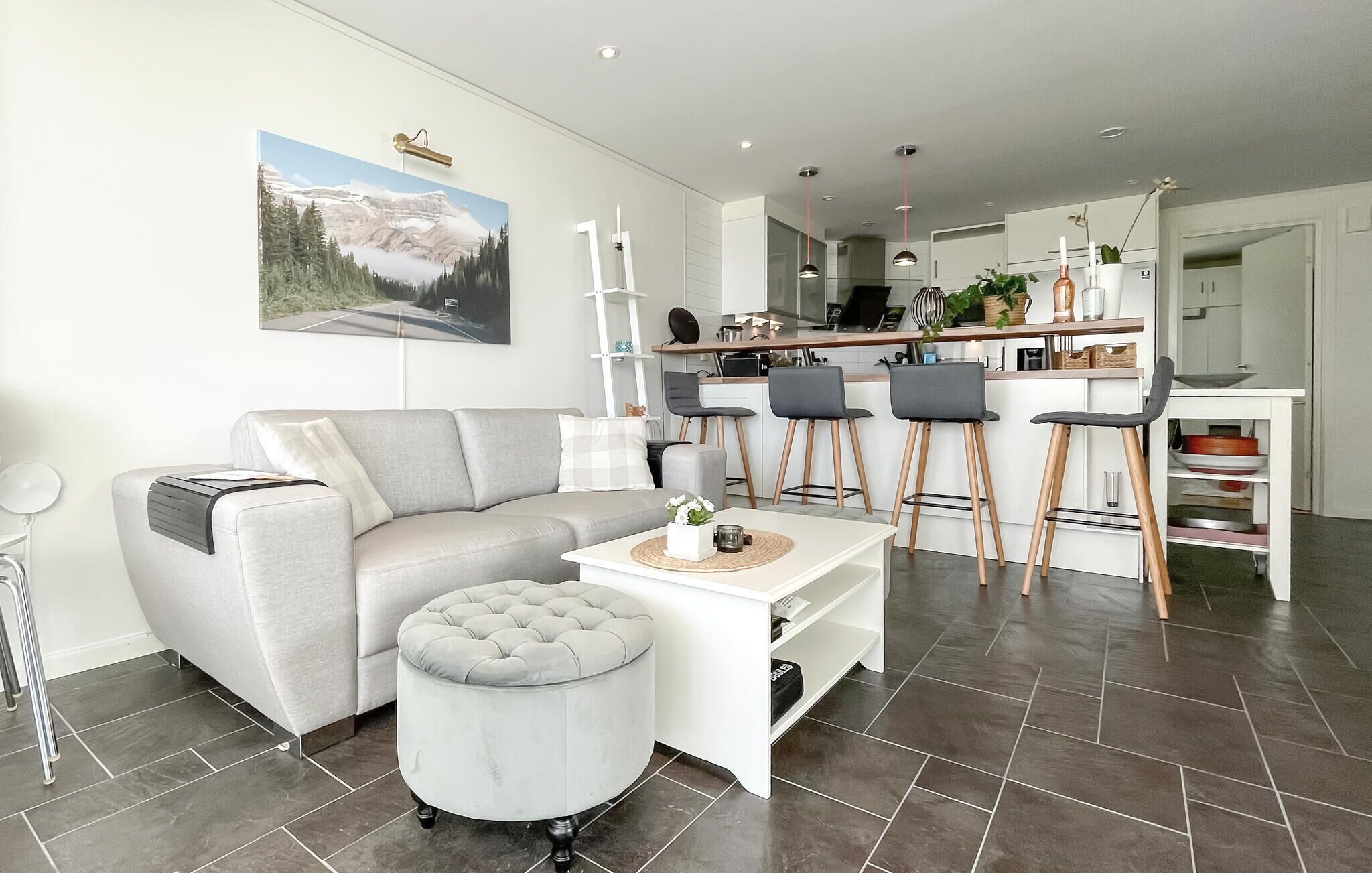 Un salon moderne avec un canapé, un pouf et une table basse. Il y a également un coin cuisine avec des tabourets de bar et une table à manger.