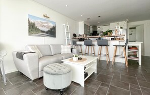 A modern living room with a sofa, ottoman, and a coffee table. There is a kitchen area with bar stools and a dining table.