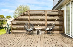 A wooden deck with two black wire chairs and a small table, a wooden slatted wall, and a potted plant.