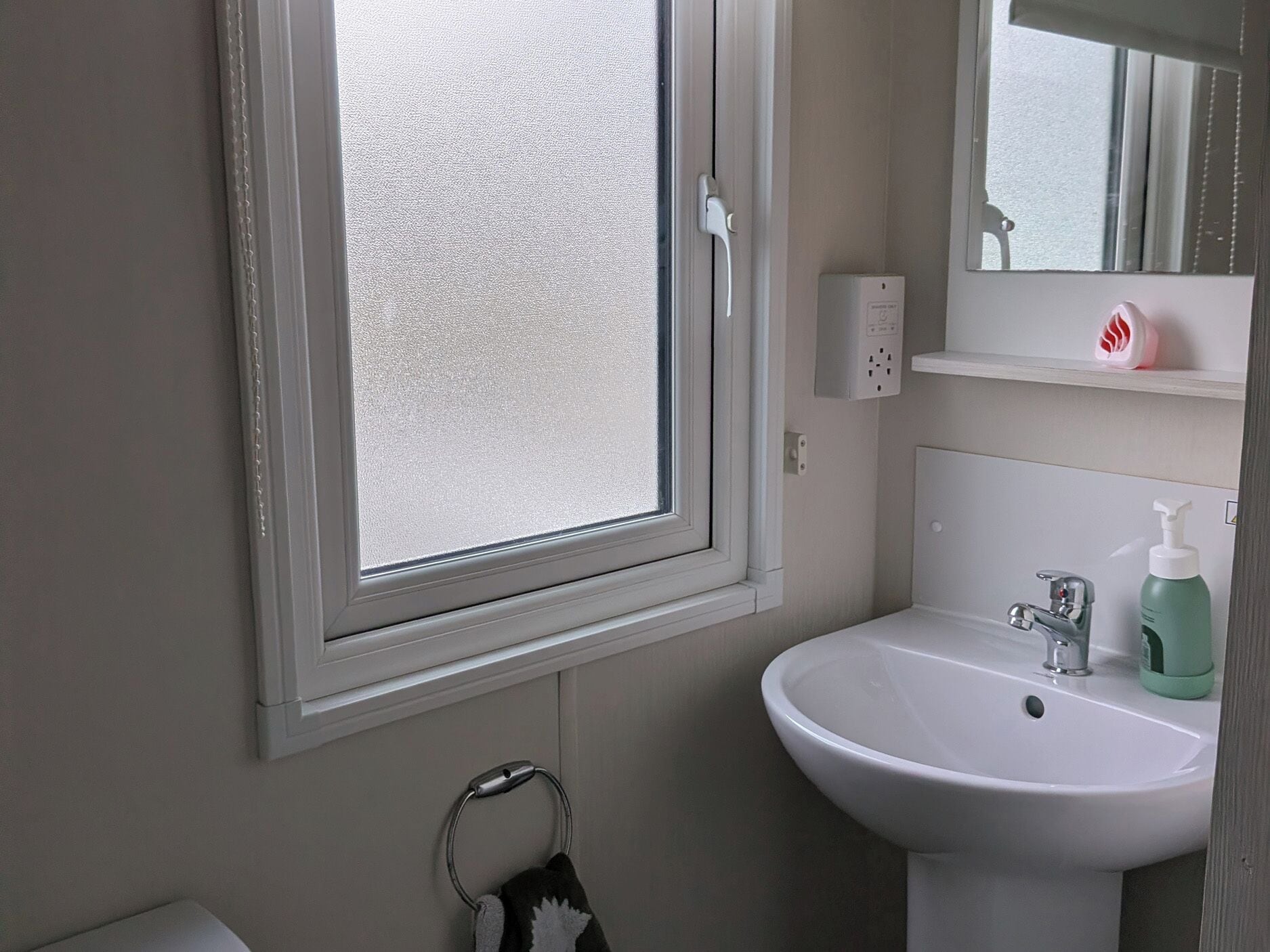 Un baño con ventana, lavamanos, espejo y perchero para toallas.
