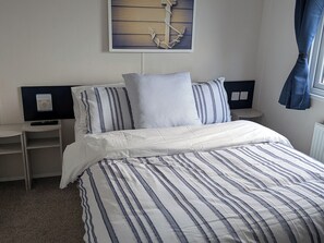 A neatly made bed with striped bedding, a bedside table with a remote control, and a wall-mounted artwork. - Presthaven - Denbigh Dunes (Holywell)