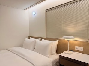 Una habitación de hotel con una cama, una mesita de noche con lámpara, un enchufe y una ventana con persianas.