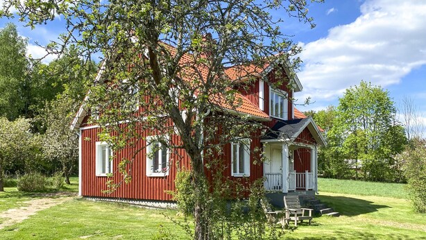 Una casa di legno rossa con un portico e due finestre, immersa nel verde.