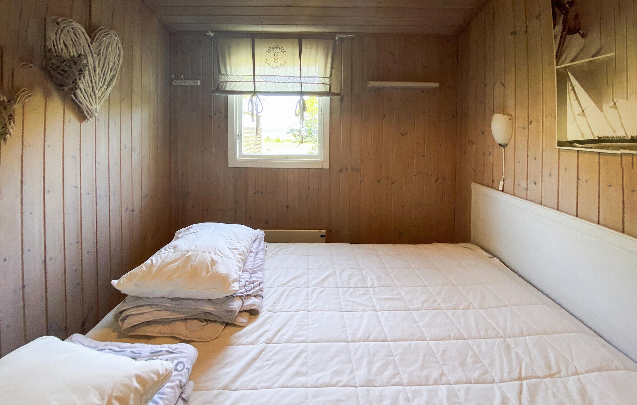 A small, cozy room with wooden walls, a single bed with white bedding, a window with a curtain, and a wall-mounted shelf with a picture.