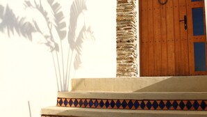A set of steps leading to a wooden door with a stone pillar, a tiled border, and a shadow of palm trees. - Asinerie Kuleni (Ponteilla)