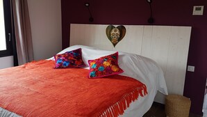 A bed with a red blanket and two colorful pillows, a white headboard, and a decorative wall piece. - Asinerie Kuleni (Ponteilla)
