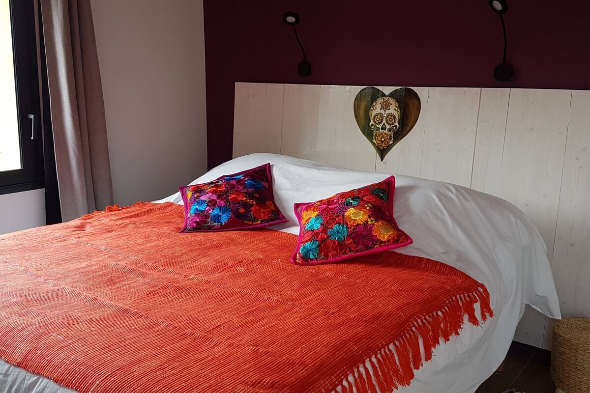 A bed with a red blanket and two colorful pillows, a white headboard, and a decorative wall piece.