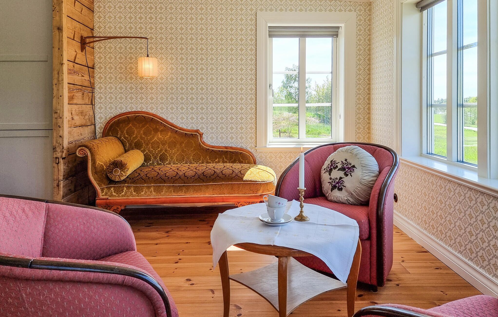 Una habitación con piso de madera, un sofá antiguo, un sillón rojo, una pequeña mesa con una taza y un platillo, y una ventana con vistas a la vegetación.