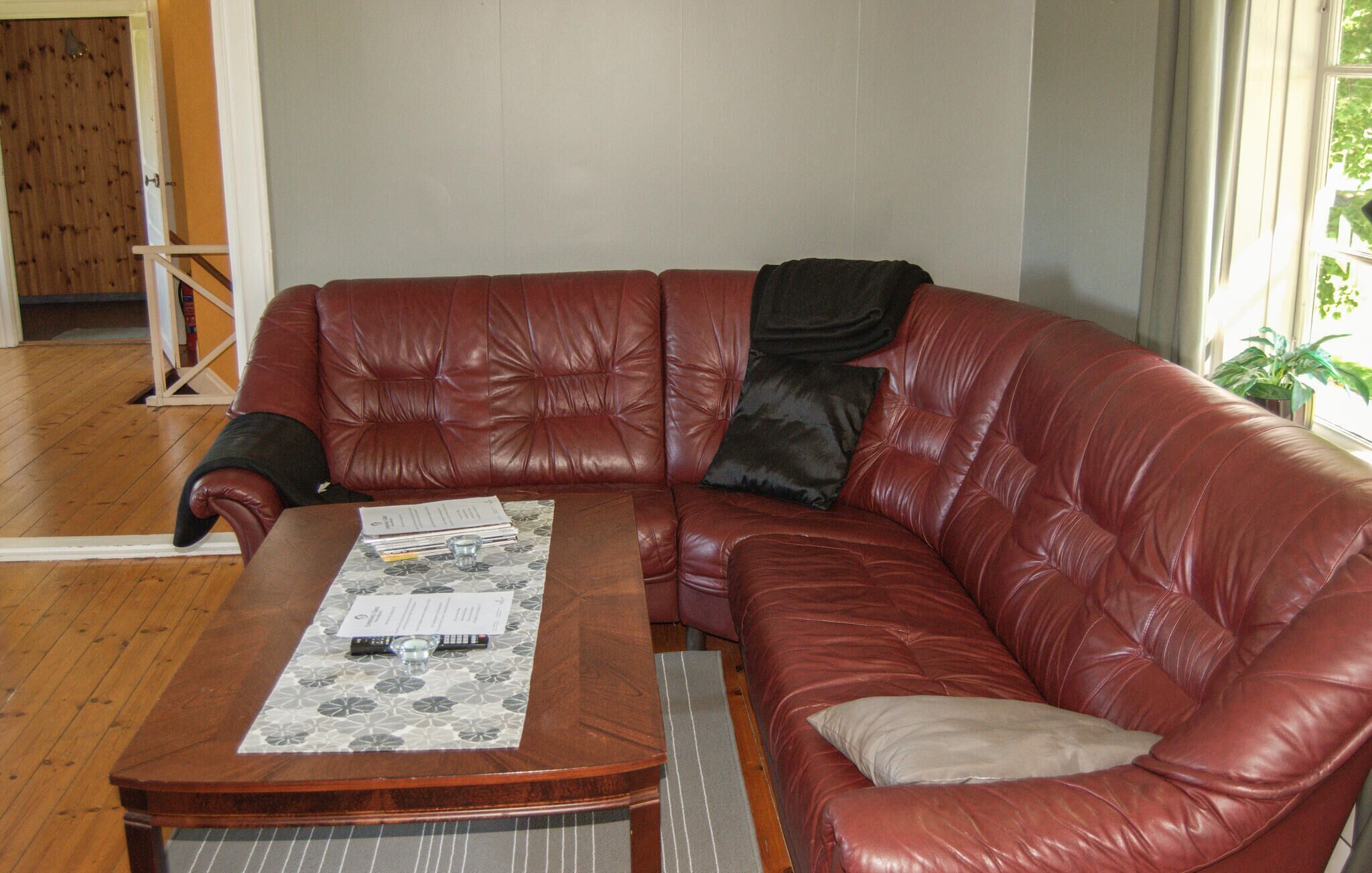 A red leather sofa with a black pillow, a wooden coffee table with papers and a glass, and a window with a plant.