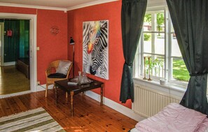 Une pièce avec un plancher en bois, un tableau représentant une zèbre accroché au mur, une chaise, une table sur laquelle se trouvent divers objets, et une fenêtre avec des rideaux.