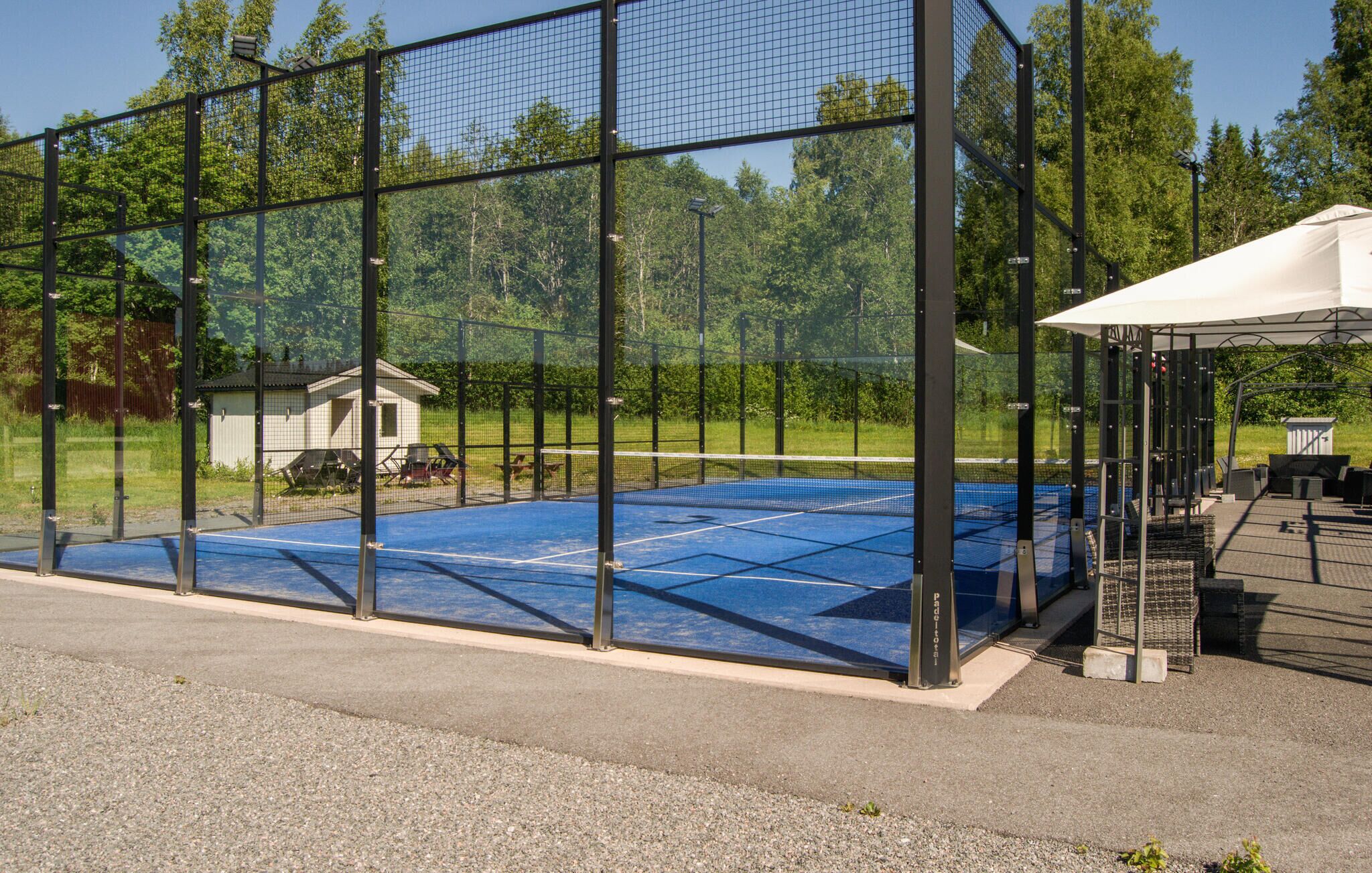 A tennis court enclosed by a netted fence, with a pavilion and a small shed in the background.