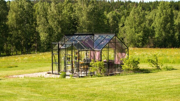 A glass greenhouse structure with a table and chairs inside, surrounded by a grassy field and a forest in the background. - 4 bedroom awesome home in Kopparberg (Kopparberg)