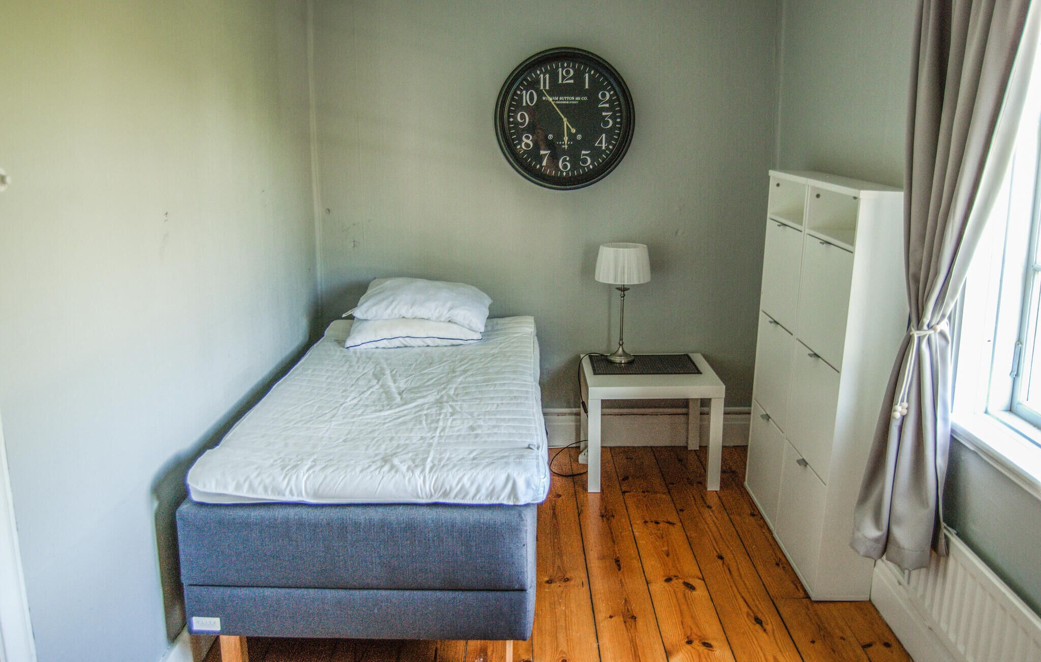 A small, simple room with a bed, a nightstand with a lamp, a wall clock, and a dresser.