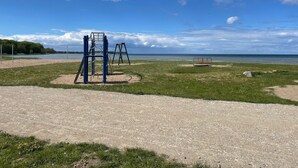 Um parquinho com estruturas de escalada e balanços, uma área com areia e um corpo d'água ao fundo.
