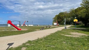 Um parque com um parquinho vermelho, uma cesta de basquete amarela, um banco e um caminho que leva à água.