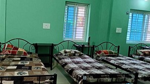A room with four single beds, each with a patterned bedspread and pillows. There are two windows with bars, one on each wall, and a door on the left. - APARNA HOTEL DORMITORY (Bhubaneshwar)