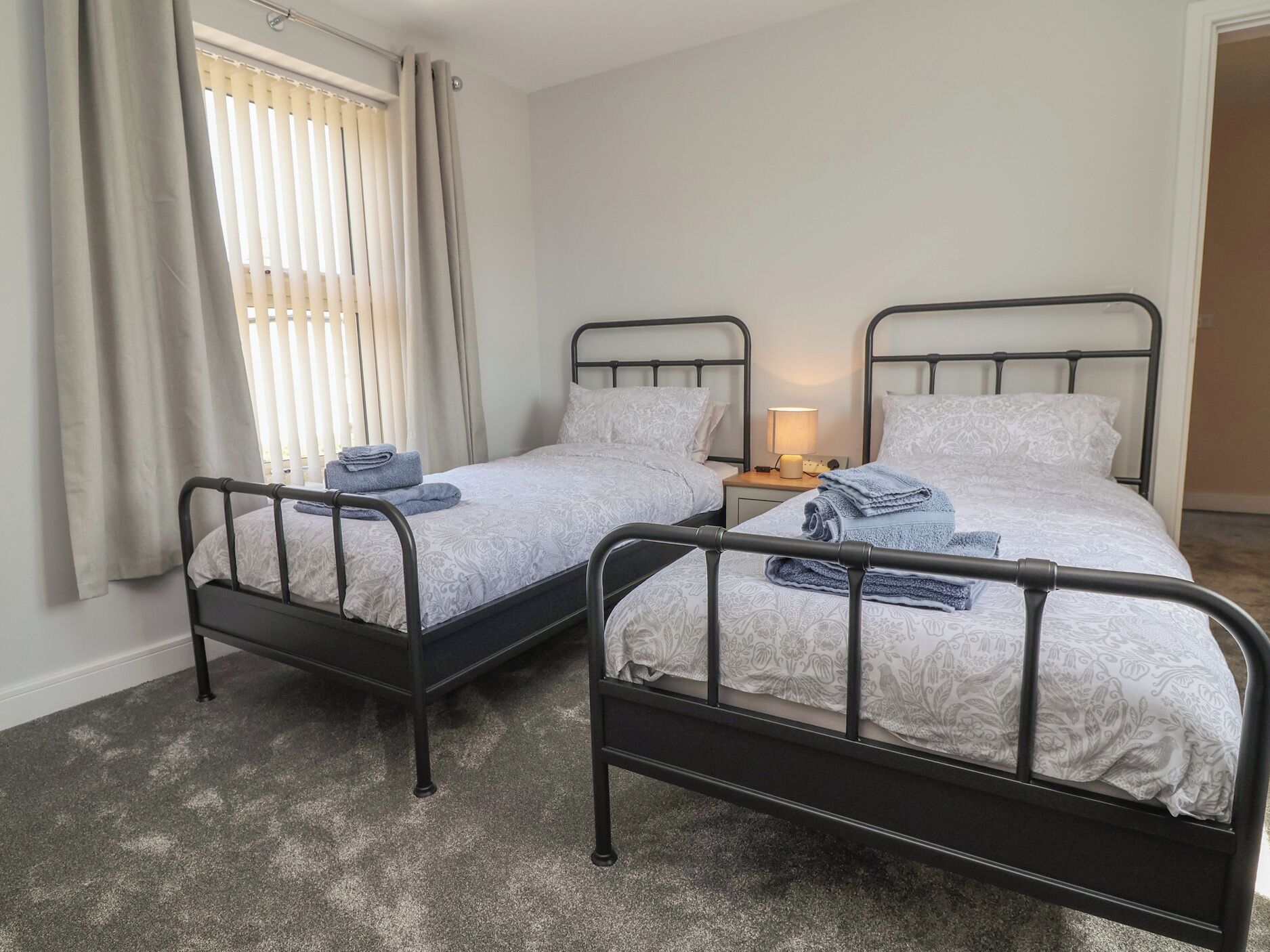 A room with two single beds, each with a metal frame, a bedside table with a lamp, and a window with blinds.