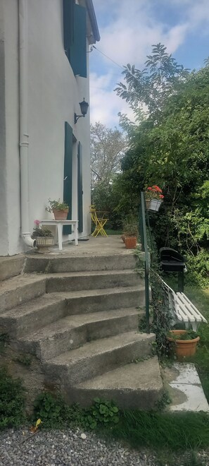 A set of concrete steps leading to a white building with green shutters, a small table with a potted plant, and a bench with a planter.