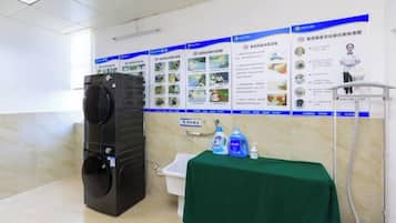 A public restroom with a large black washing machine, a sink, a green table with cleaning supplies, and informational posters on the wall.