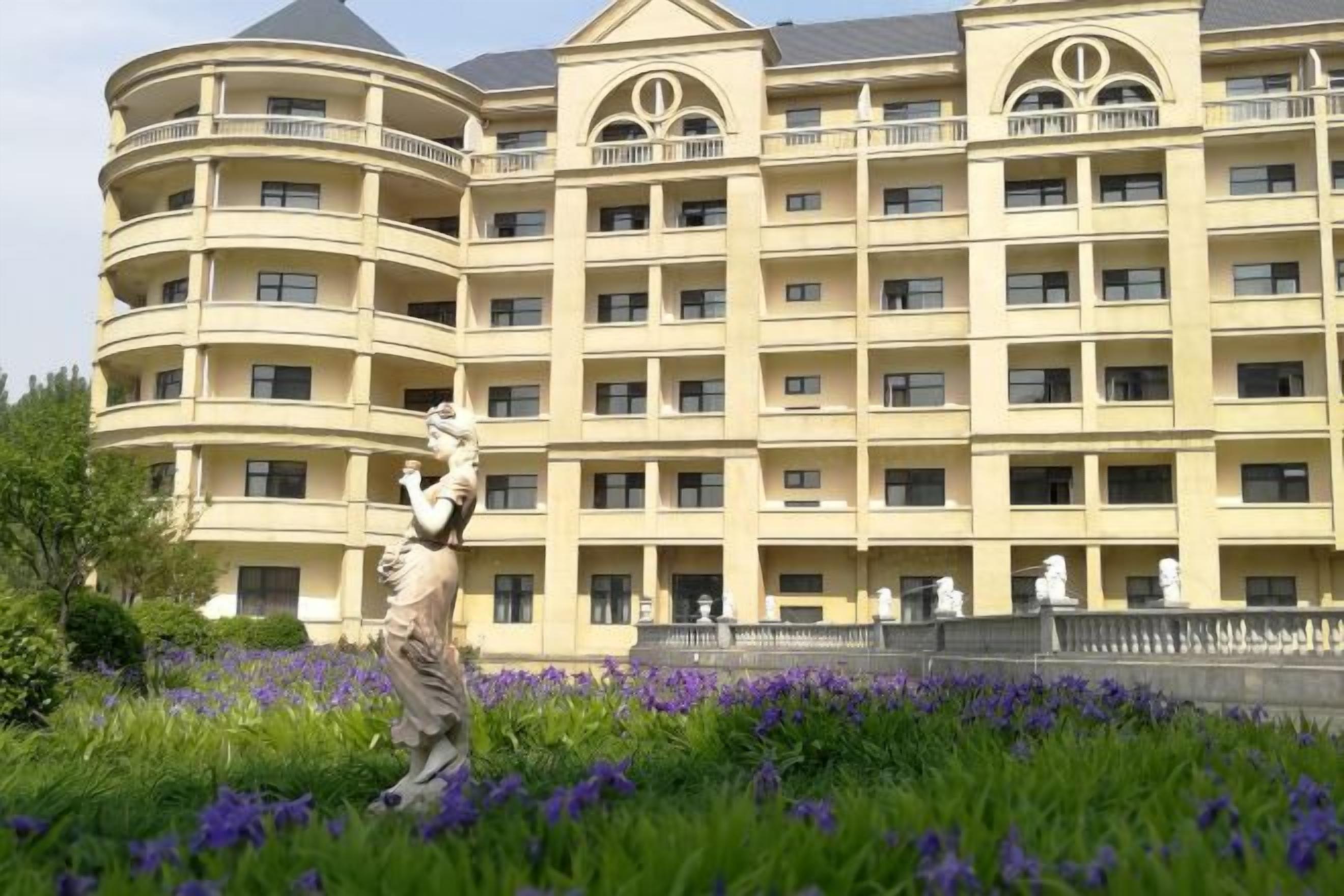 Hotel de diversos pisos amb balcons, una estàtua al primer pla i un jardí ben cuidat amb flors morades.