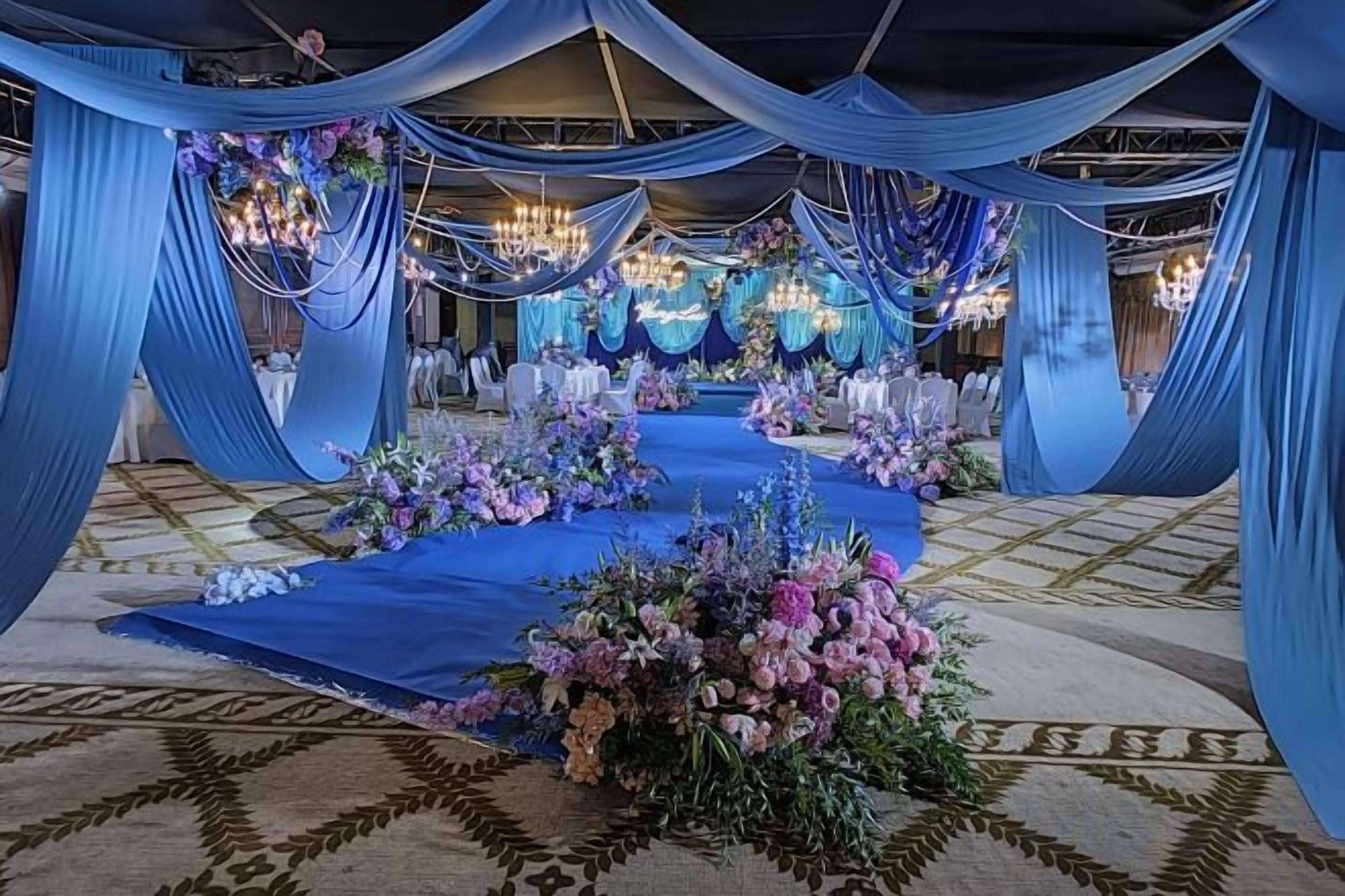 An indoor event space with draped blue fabric, floral centerpieces, and chandeliers.