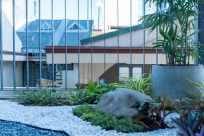 Un jardin avec des rochers et des plantes, un bac à fleurs en béton et, au fond, un bâtiment avec un toit rouge.