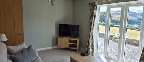 A living room with a sofa, a television on a wooden stand, a desk with a laptop, and a view of the outdoors through large windows.