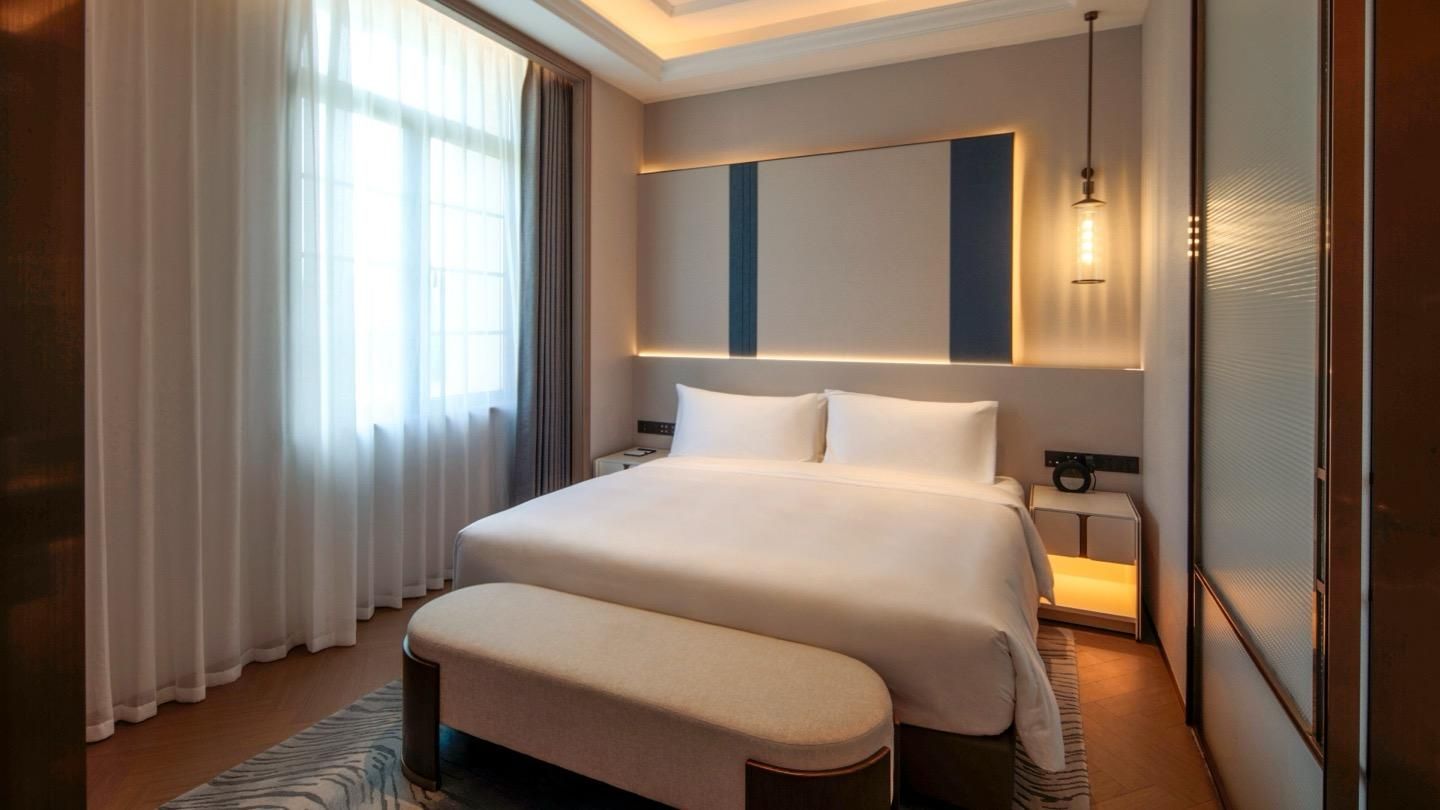 Quarto de hotel com cama grande, mesas de cabeceira, um criado-mudo, janela com cortinas e luminária moderna.