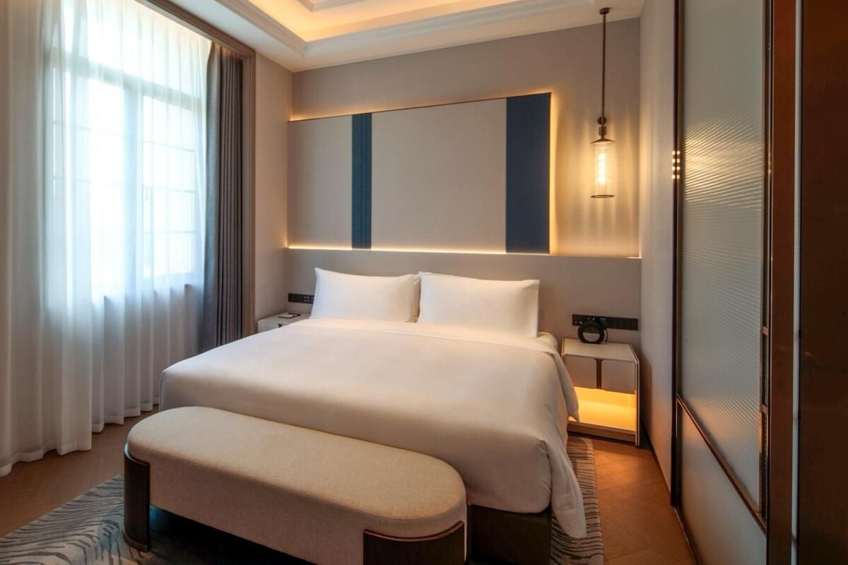 A hotel room with a large bed, bedside tables, a nightstand, a window with curtains, and a modern light fixture.