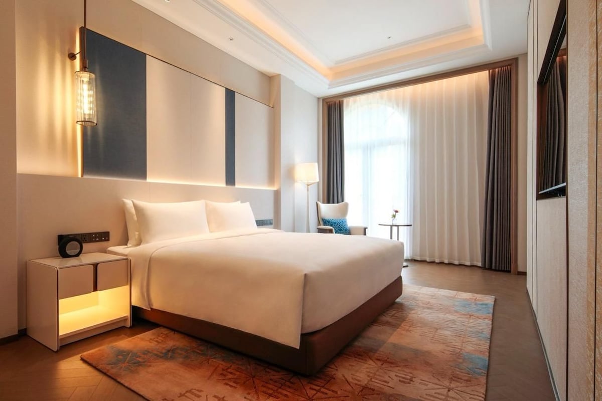 A modern hotel room with a large bed, bedside tables, a wall-mounted light, and a view of the outdoors through the window.