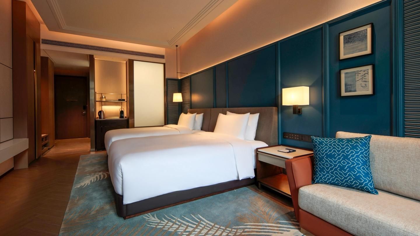A hotel room with a double bed, a sofa, a nightstand, a lamp, and artwork on the wall.